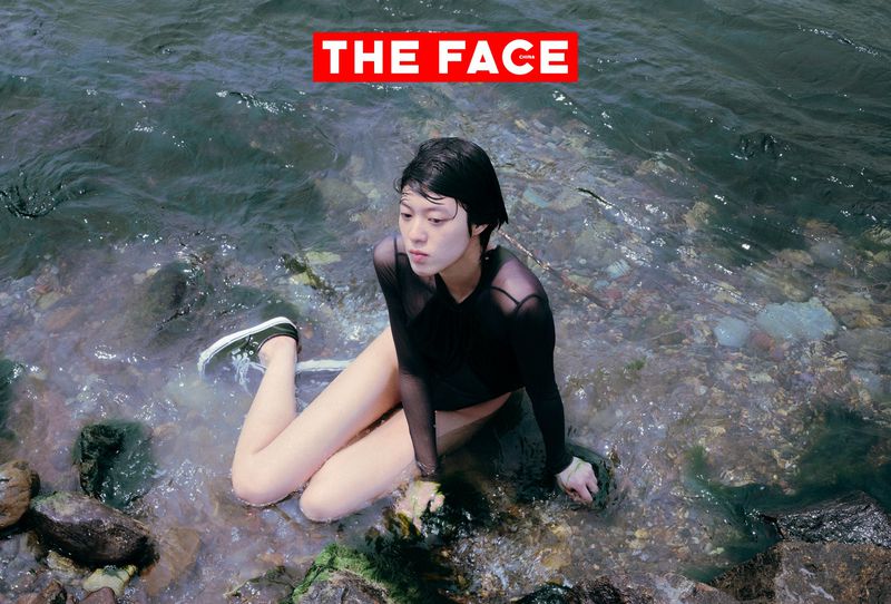East Asian model with short wet hair wearing a black mesh top sitting in shallow river water over submerged riverbed stones.