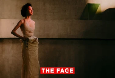 East Asian female model with short hair standing in a sculpted beige halter dress against a concrete wall under warm light.