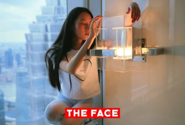 An East Asian model perched beside a high-rise window reaching toward a modern glass wall sconce with a city skyscraper view.