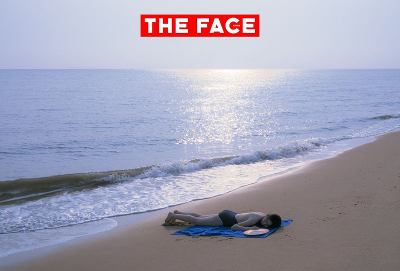 Model lying face down on a blue towel on a sandy beach near gentle ocean waves under bright daylight.