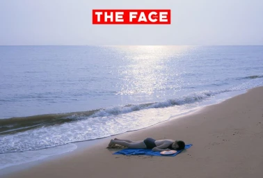 Model lying face down on a blue towel on a sandy beach near gentle ocean waves under bright daylight.