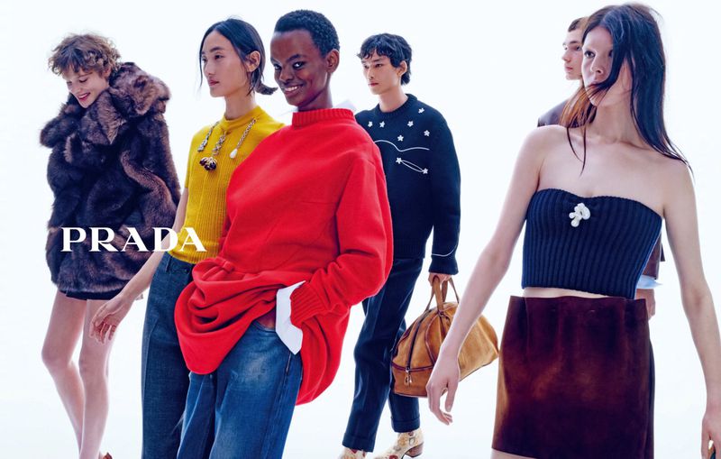 Group of fashion models walking against a bright white studio backdrop wearing red knitwear, yellow top, and fur coat.