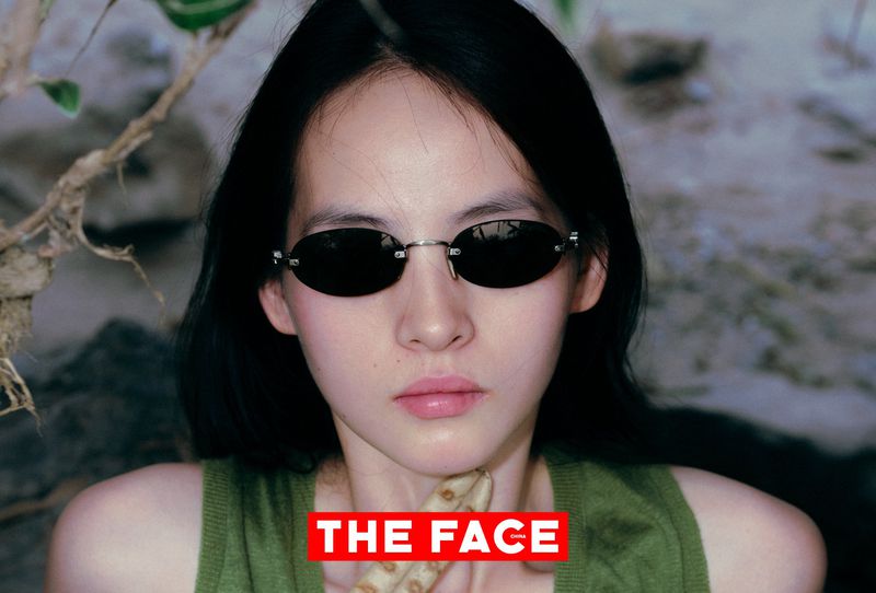 Close-up portrait of an East Asian model wearing small oval sunglasses and an olive green sleeveless top outdoors.