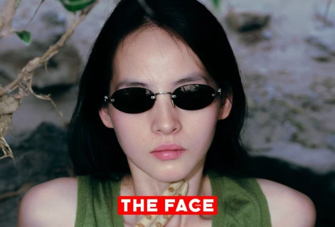 Close-up portrait of an East Asian model wearing small oval sunglasses and an olive green sleeveless top outdoors.