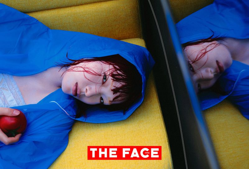 Model in a vivid blue hooded cape lying down on a yellow textured sofa holding a red tomato next to her mirror reflection.