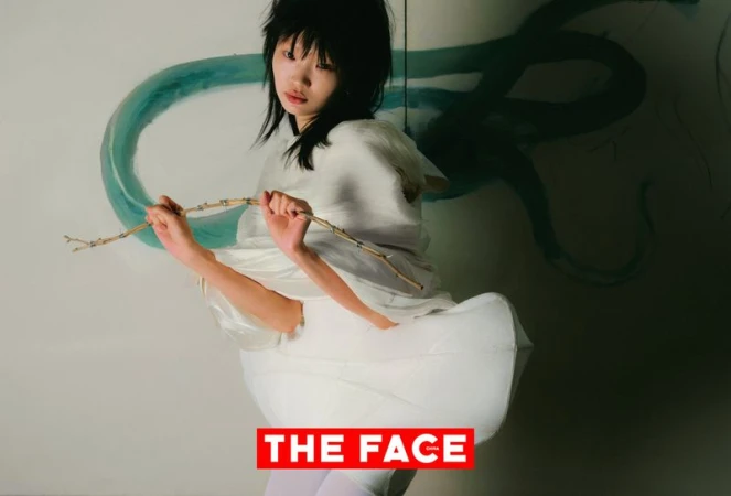 East Asian model with shaggy dark hair wearing a sculptural white dress and holding a wooden branch against a painted wall.