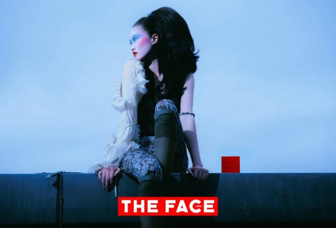 Model with voluminous bouffant hair and bold blue eye makeup seated on a ledge against a pale blue sky.