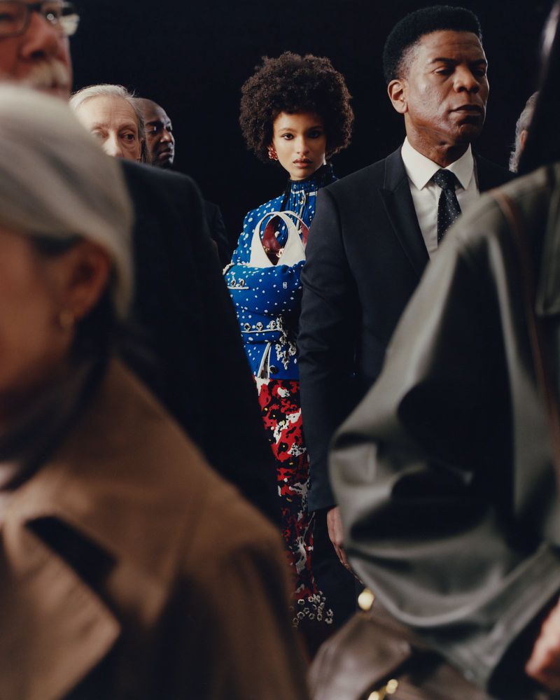 Model Ana Portela in a blue patterned top stands out surrounded by a crowd in dark formal attire.