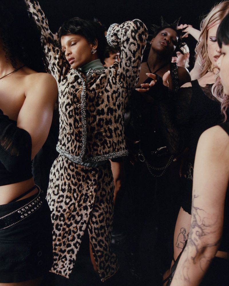 Model posing in a leopard print tweed skirt suit surrounded by a crowd of models dressed in black against a dark studio background.