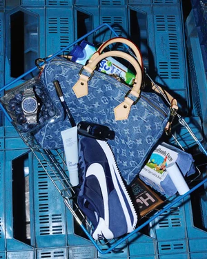 Top-down view of a wire shopping basket filled with a denim monogram bag, navy sneaker, luxury watch, and groceries over blue crates.