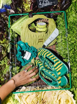 Top-down view of a green shopping basket filled with a lime handbag, striped shoes, and beauty products surrounded by fresh produce.