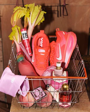 Wire shopping basket filled with pink sneakers, rhubarb stalks, cosmetics, perfume, and peaches for British Vogue.