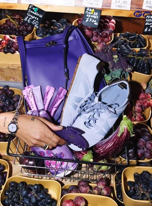 A wire shopping basket holding a purple handbag, lilac sneakers, and an eggplant surrounded by crates of dark purple grapes.