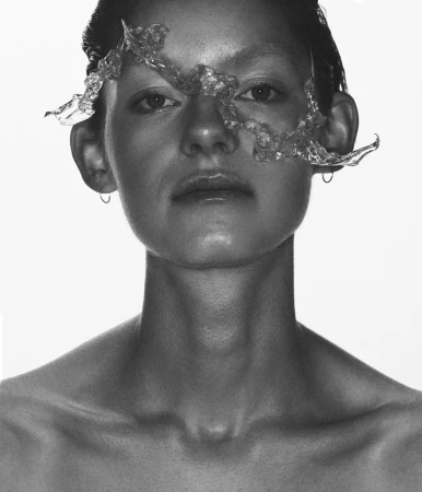 Monochrome close-up portrait of a woman wearing an abstract clear sculptural mask across her eyes on a bright white background.