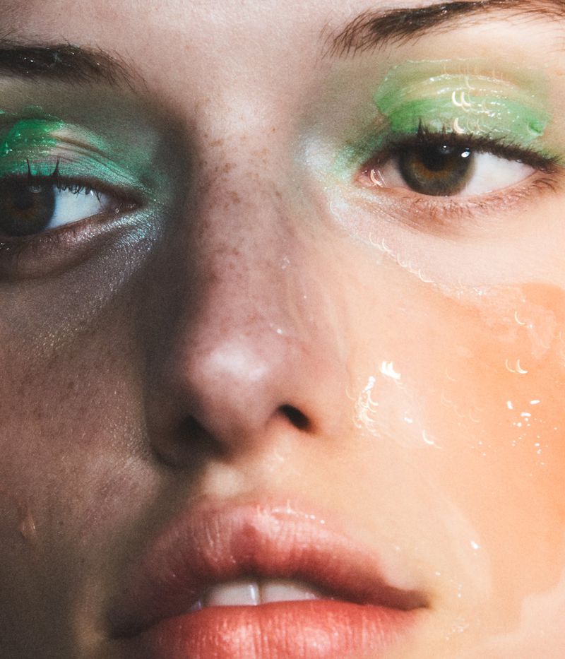 Close-up beauty portrait of a woman with freckled skin, green glossy eye makeup, and dewy wet-look textures.