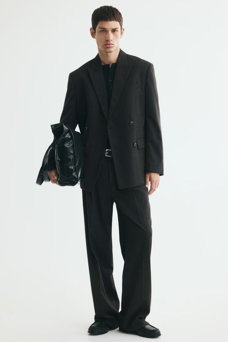 Model Luka Tesic wearing an oversized black double-breasted suit and holding a black leather bag against a white studio background.