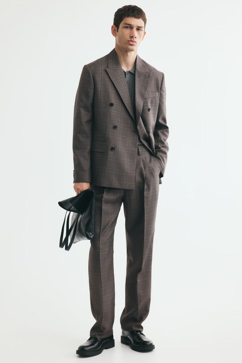Model Luka Tesic posing in a brown plaid double-breasted suit and holding a black tote bag against a white studio background.