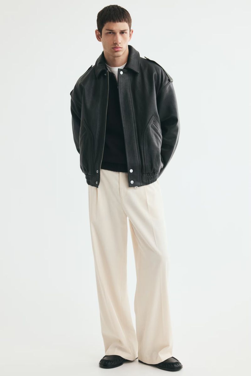 Model Luka Tesic wearing a black leather bomber jacket and wide-leg cream trousers against a white seamless backdrop.
