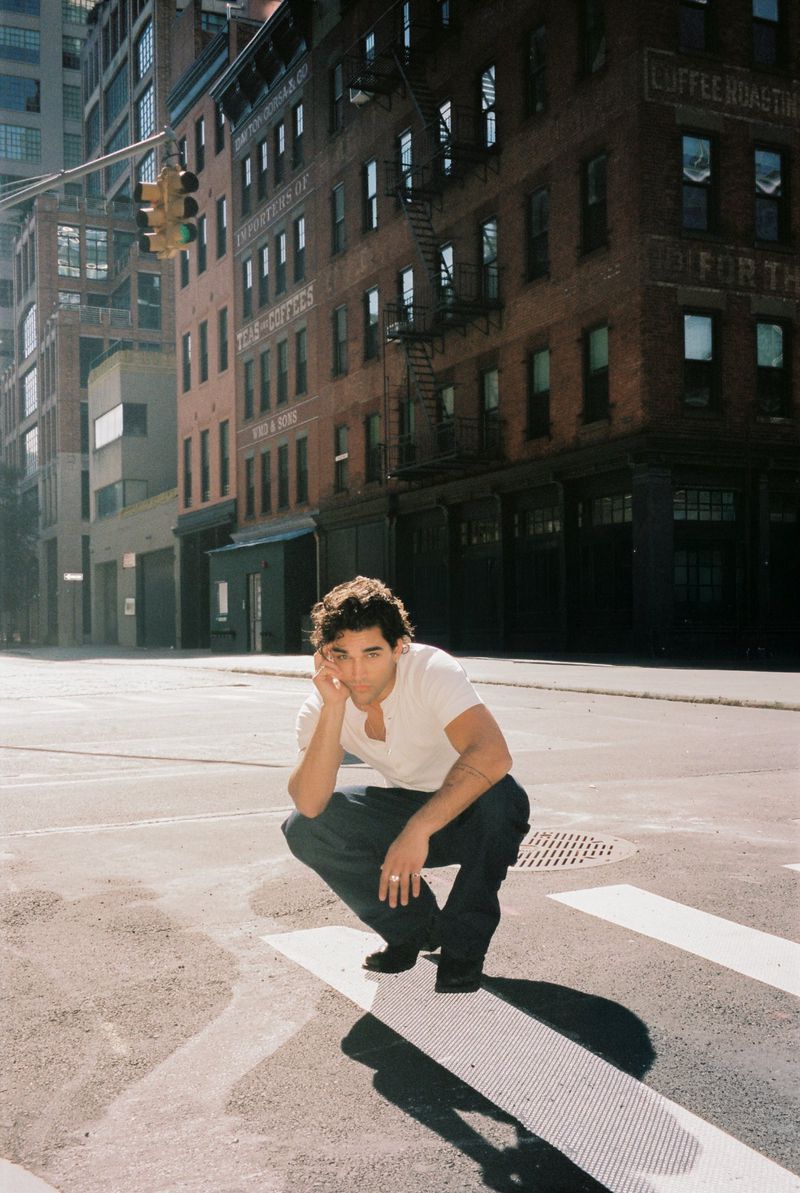 Man crouching on an asphalt crosswalk wearing a white t-shirt and dark blue denim jeans in a city street setting.