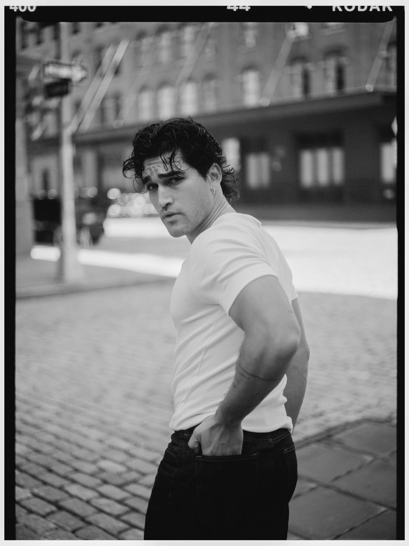 Man in a white t-shirt and dark jeans looking over his shoulder on a cobblestone city street in black and white film.