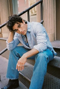 Man with dark curly hair wearing a striped shirt and blue jeans sitting on outdoor metal steps.