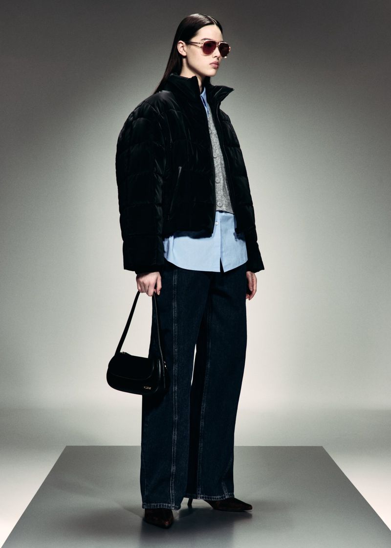 Fashion model wearing a black puffer jacket, wide-leg denim jeans, sunglasses, and carrying a black shoulder bag in a studio.