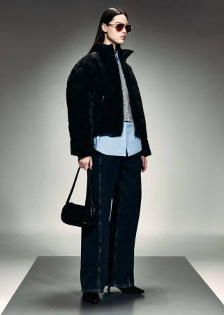 Fashion model wearing a black puffer jacket, wide-leg denim jeans, sunglasses, and carrying a black shoulder bag in a studio.