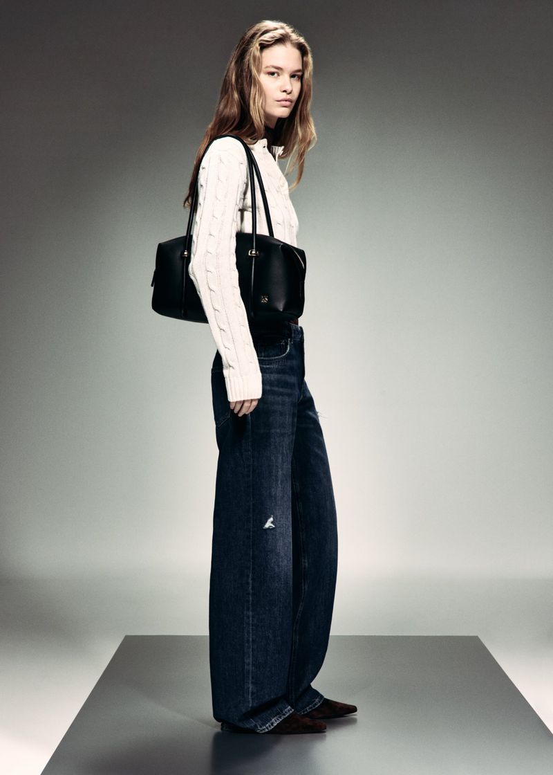 A woman poses in a cream cable-knit sweater, wide-leg dark wash jeans, and a black shoulder bag on a studio platform.