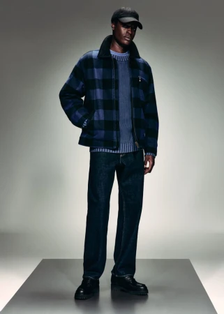 Black male model in a blue and black plaid zip jacket, dark denim jeans, black baseball cap, and black leather shoes on a platform.