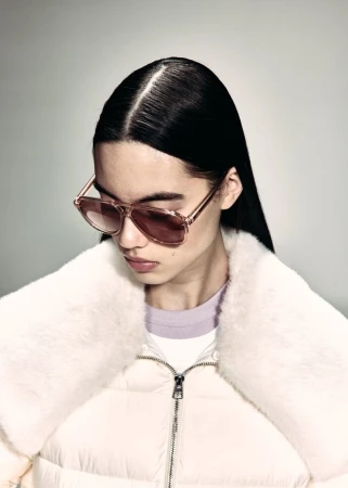 Model wearing pink-tinted sunglasses and a white shearling-collar puffer jacket looking down against a pale studio backdrop.