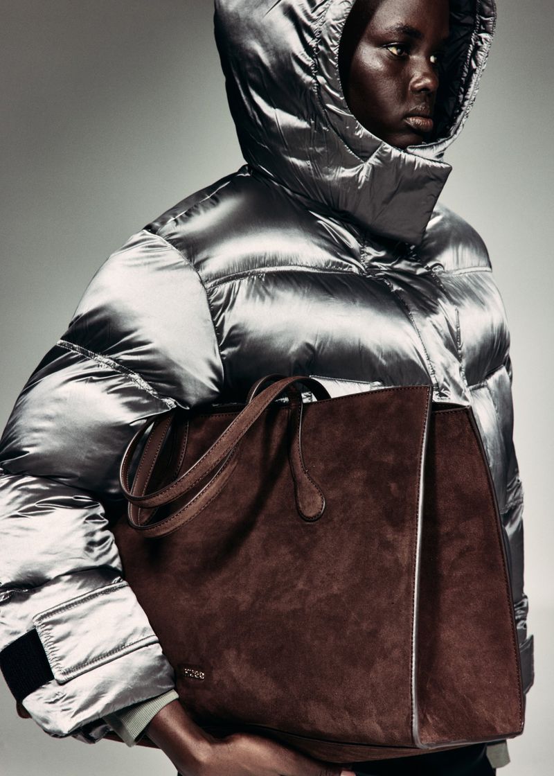 Model wearing a shiny metallic silver hooded puffer jacket while carrying a large brown suede tote bag against a studio backdrop.