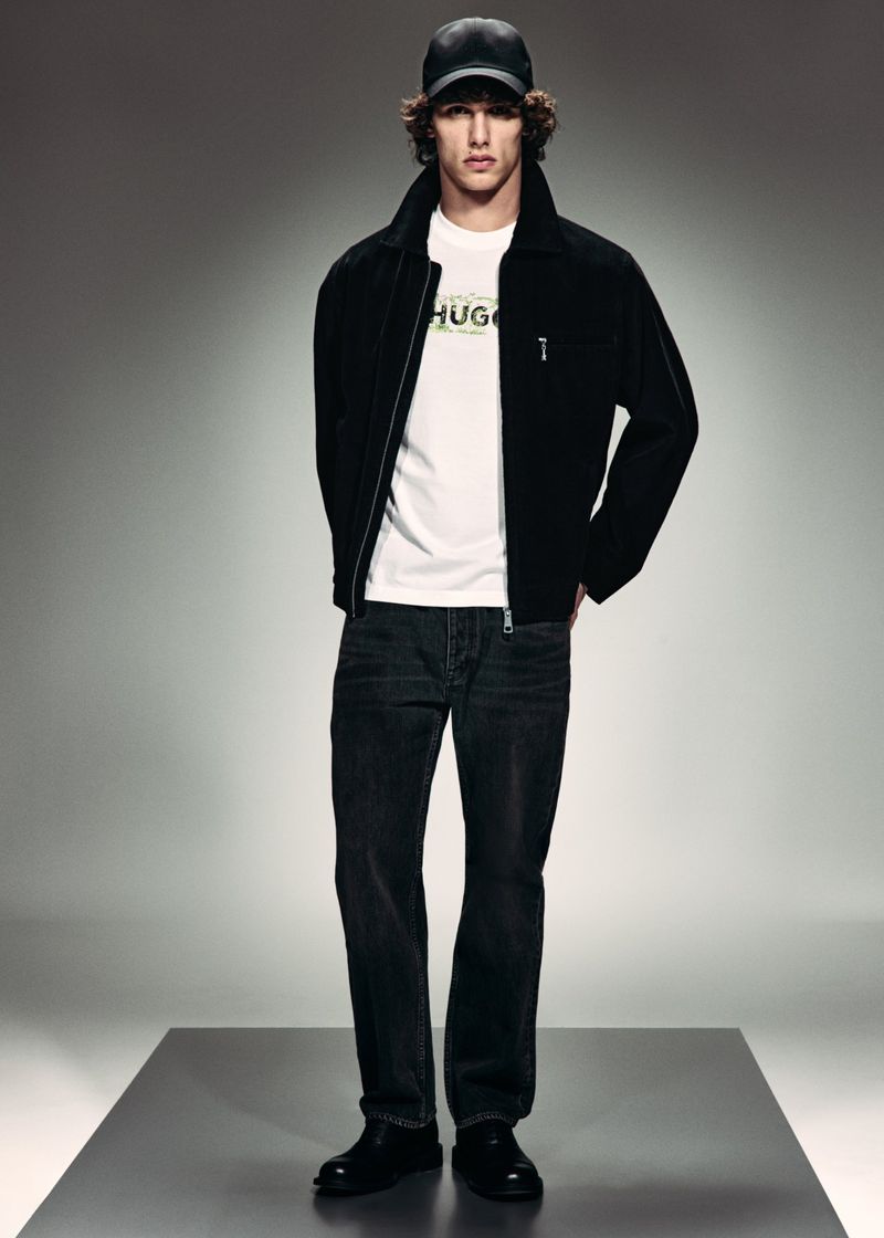Model wearing a black jacket, white HUGO logo t-shirt, dark jeans, and a black baseball cap standing on a grey studio platform.