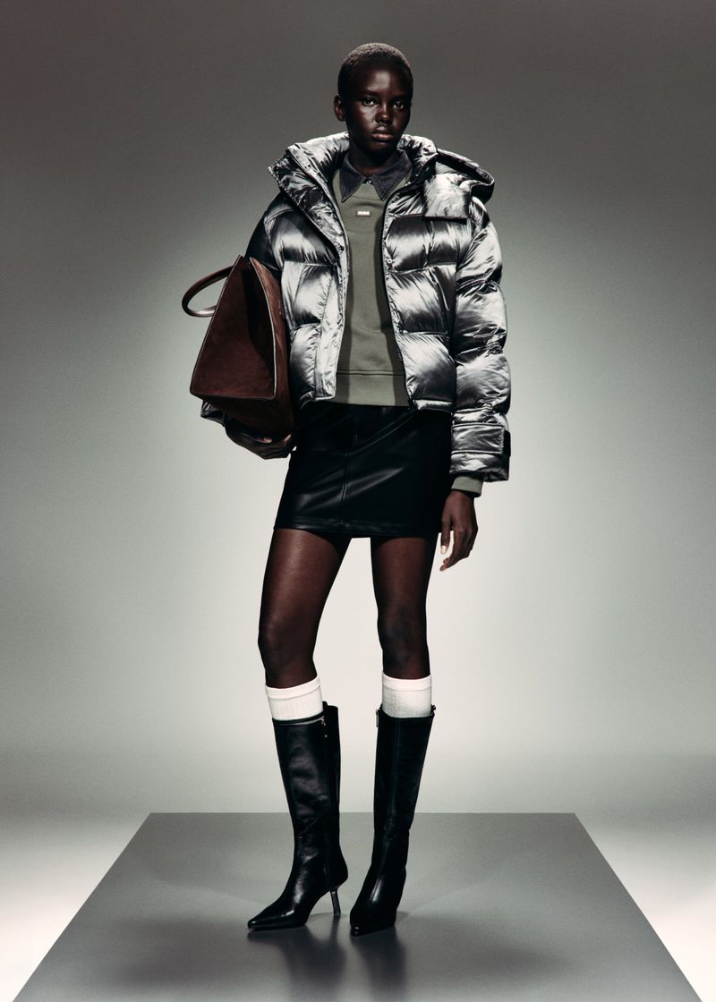A model in a metallic silver puffer jacket, black mini skirt, and knee-high leather boots holding a brown bag on a grey platform.