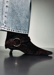 Close-up of a dark brown suede kitten heel ankle boot with a silver ring buckle strap, worn with light-wash denim jeans.