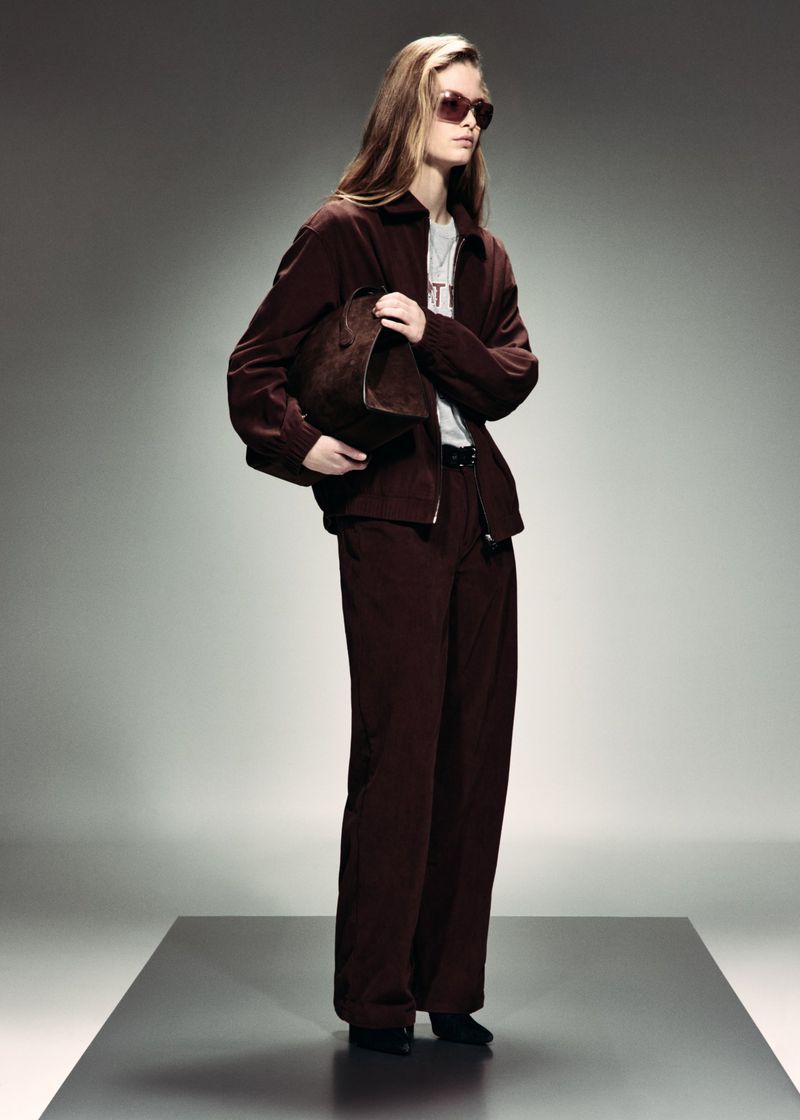 Model wearing dark brown jacket and matching wide-leg trousers with sunglasses, holding a clutch on a studio platform.