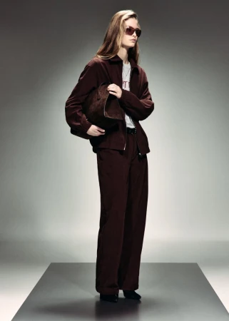 Model wearing dark brown jacket and matching wide-leg trousers with sunglasses, holding a clutch on a studio platform.