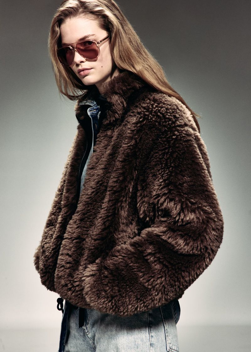 Model wearing tinted aviator sunglasses and a plush brown faux fur jacket with denim against a grey studio backdrop.