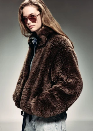 Model wearing tinted aviator sunglasses and a plush brown faux fur jacket with denim against a grey studio backdrop.