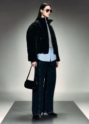 Model wearing a black puffer jacket, layered light blue shirt, wide-leg dark jeans, sunglasses, and holding a black shoulder bag in studio.