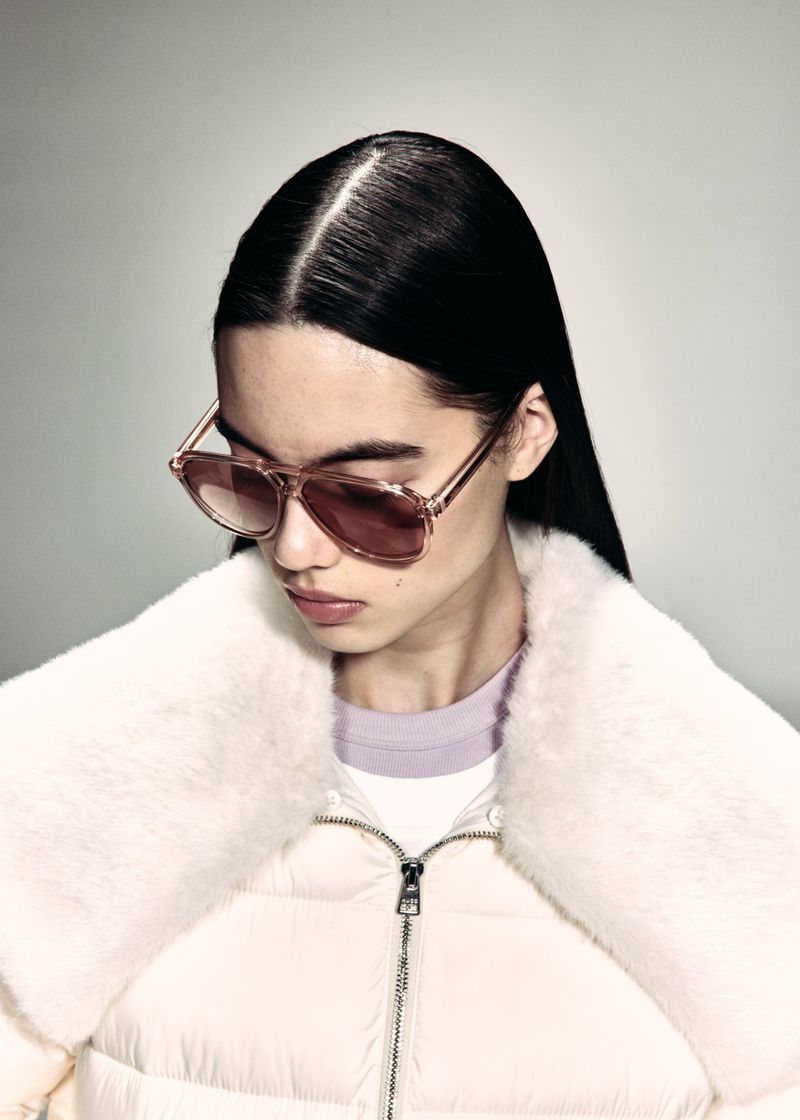 Model wearing tinted aviator sunglasses and an off-white puffer jacket with a plush faux fur collar against a grey studio backdrop.