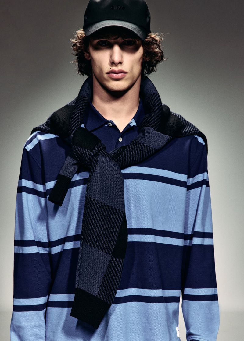 Male model wearing a black cap, striped blue rugby polo, and tied knit sweater against a neutral grey studio backdrop.