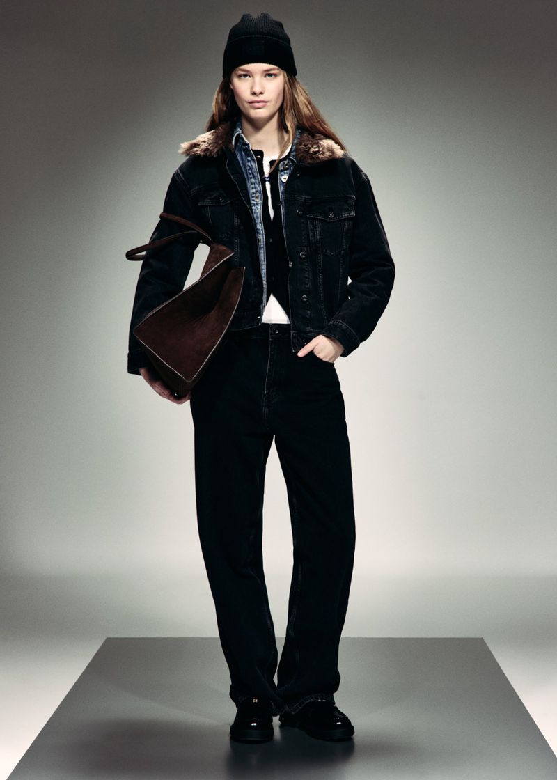 Model wearing a black shearling-collar denim jacket, black beanie, and black jeans, holding a brown suede bag in a studio.