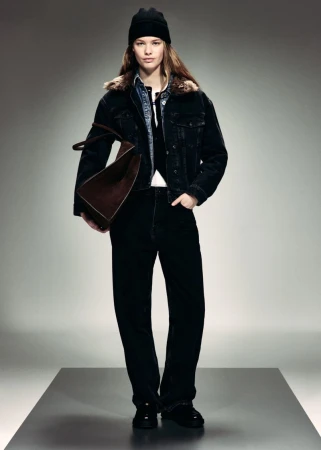 Model wearing a black shearling-collar denim jacket, black beanie, and black jeans, holding a brown suede bag in a studio.