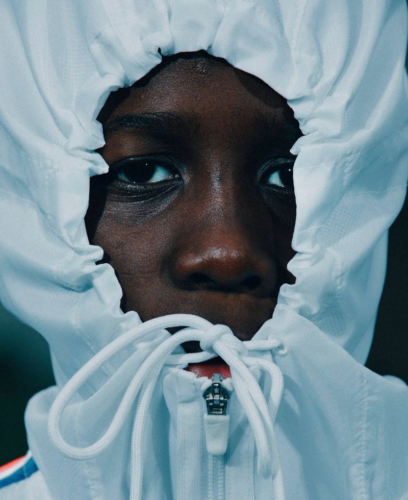 Extreme close-up portrait of a dark-skinned person whose face is tightly framed by a white hooded jacket with cinched and tied drawstrings.