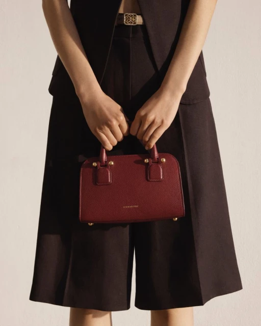 Burgundy handbag held by a person wearing a black vest and culottes, accessorized with a patterned belt, against an off-white background.
