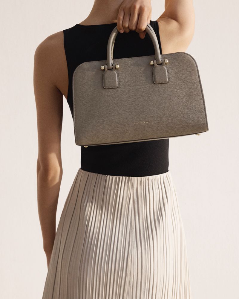 Woman holds a grey satchel handbag, wearing a black top and pleated skirt in a studio shot emphasizing fashion and accessory.