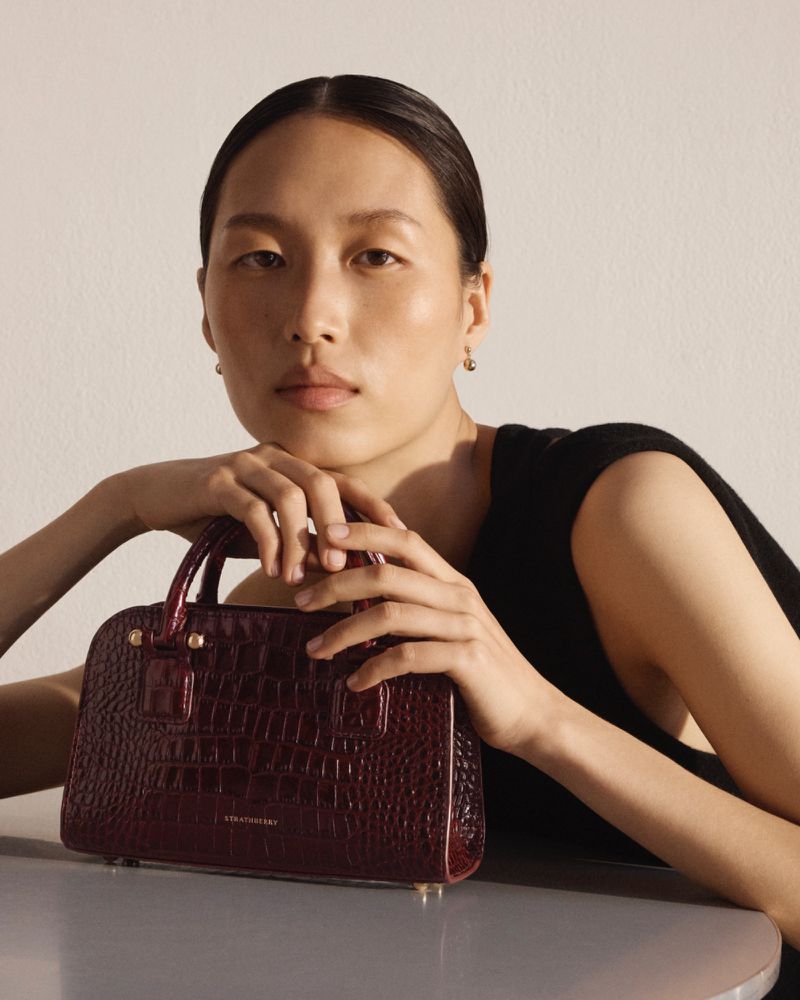 Glamorous portrait of an Asian woman showcasing a burgundy crocodile-textured handbag in a close-up, indoor setting with soft lighting.