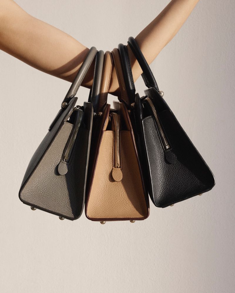 Close-up of a person holding three structured leather handbags in gray, tan, and black against a soft white background. Fashion product shot.