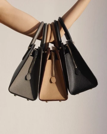 Close-up of a person holding three structured leather handbags in gray, tan, and black against a soft white background. Fashion product shot.