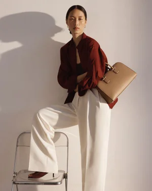 Model Yuanyuan Zhang posing in a burgundy shirt and white trousers against a white wall for Strathberry Georgia Campaign